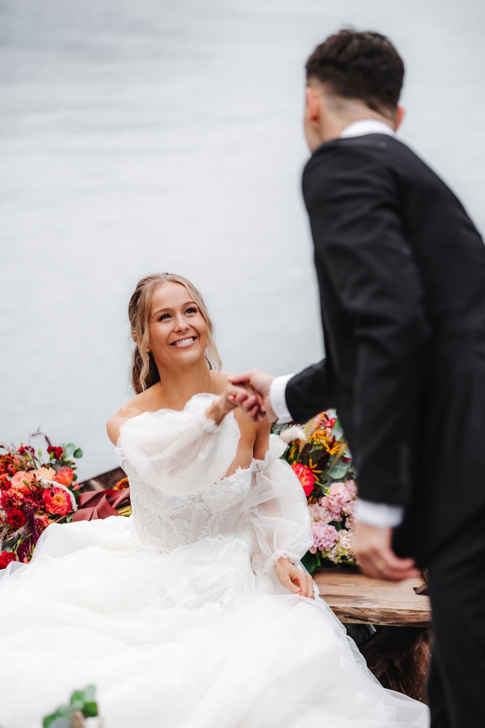 A bride in a flowing white dress smiles while sitting beside colorful floral arrangements at an outdoor wedding venue.