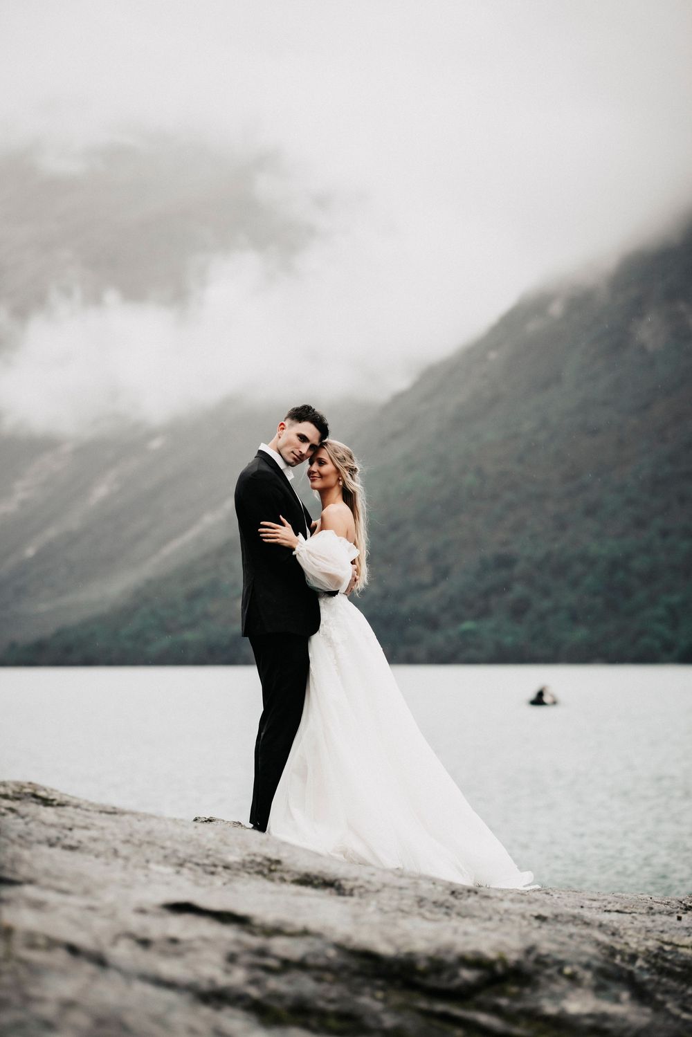A bride and groom embrace on a rocky shore with misty mountains and a lake in the background.