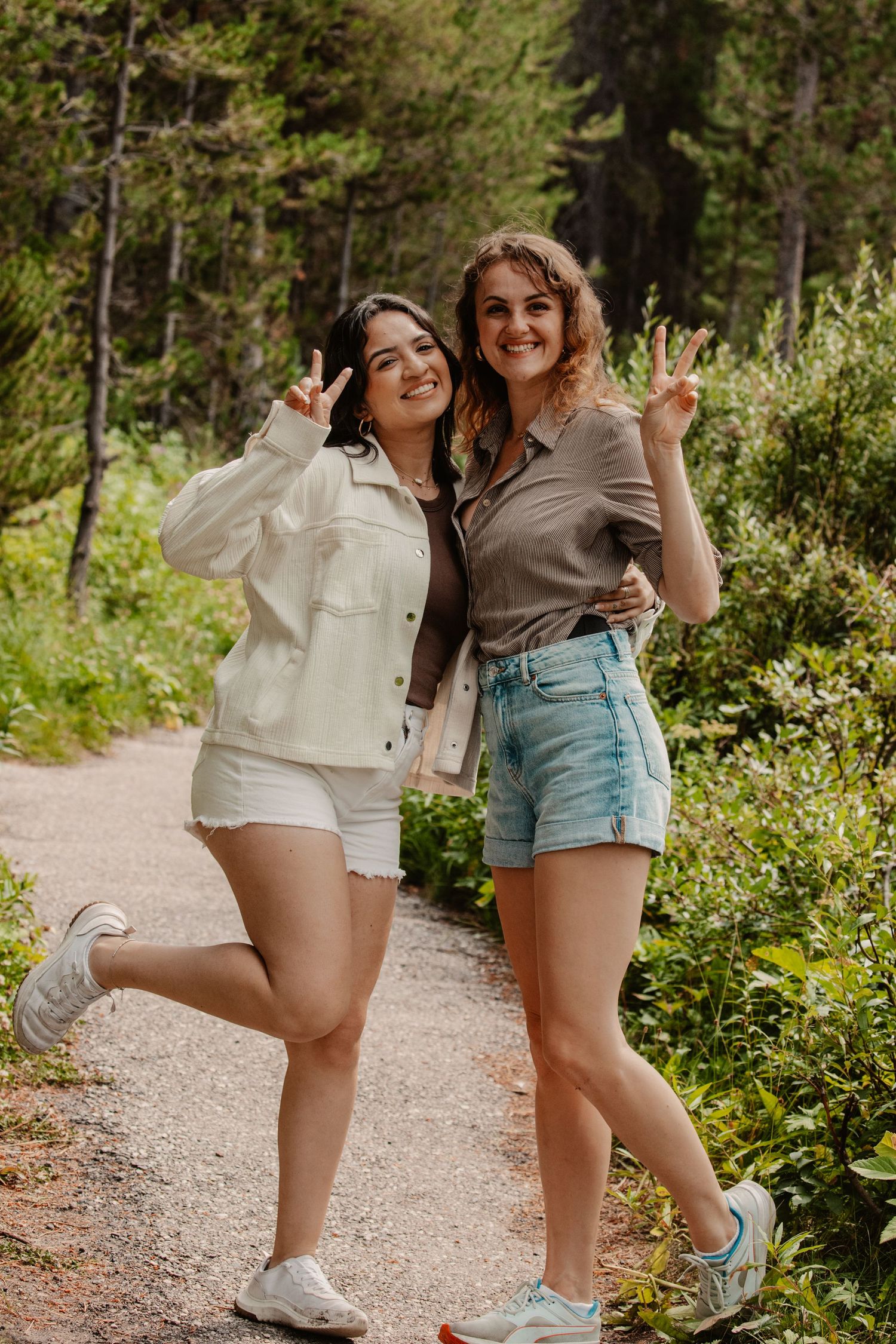 Two friends in casual summer outfits posing playfully on forest hiking trail.