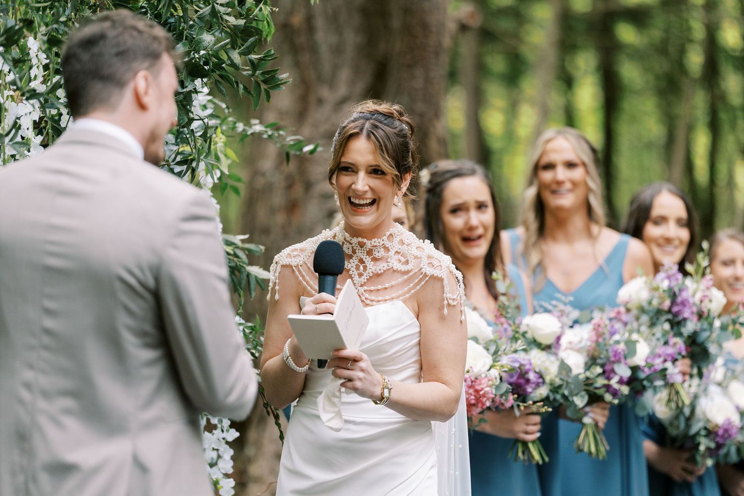 Couple exchanges vows during an outdoor wedding ceremony while bridesmaids in blue dresses hold purple bouquets at Windham Manor.