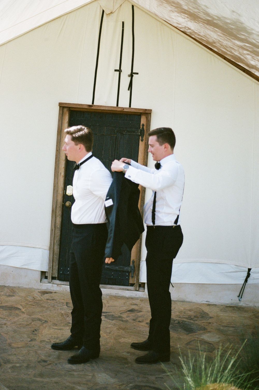 Groomsmen helping with formal attire preparations in white tent before ceremony.