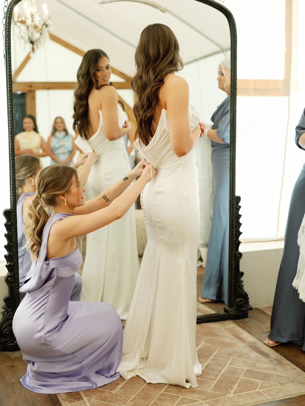 Bride getting help with wedding dress adjustments while looking in a full-length mirror.