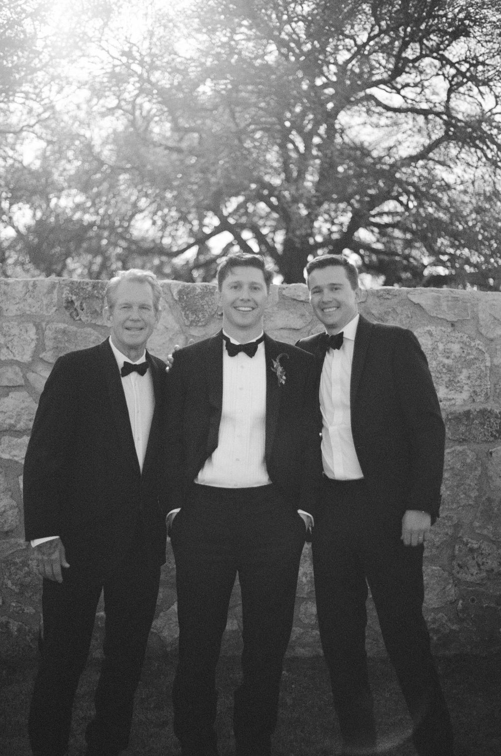 Three men in tuxedos pose together against a stone wall backdrop.