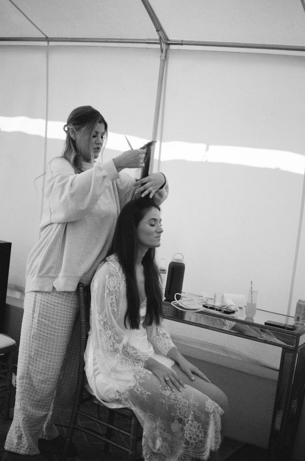 Wedding preparations show a stylist working on a bride's hair in a white tent setting.