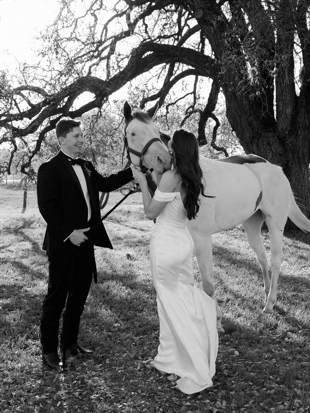 A couple in wedding attire poses with a white horse under a large tree in a rustic setting.