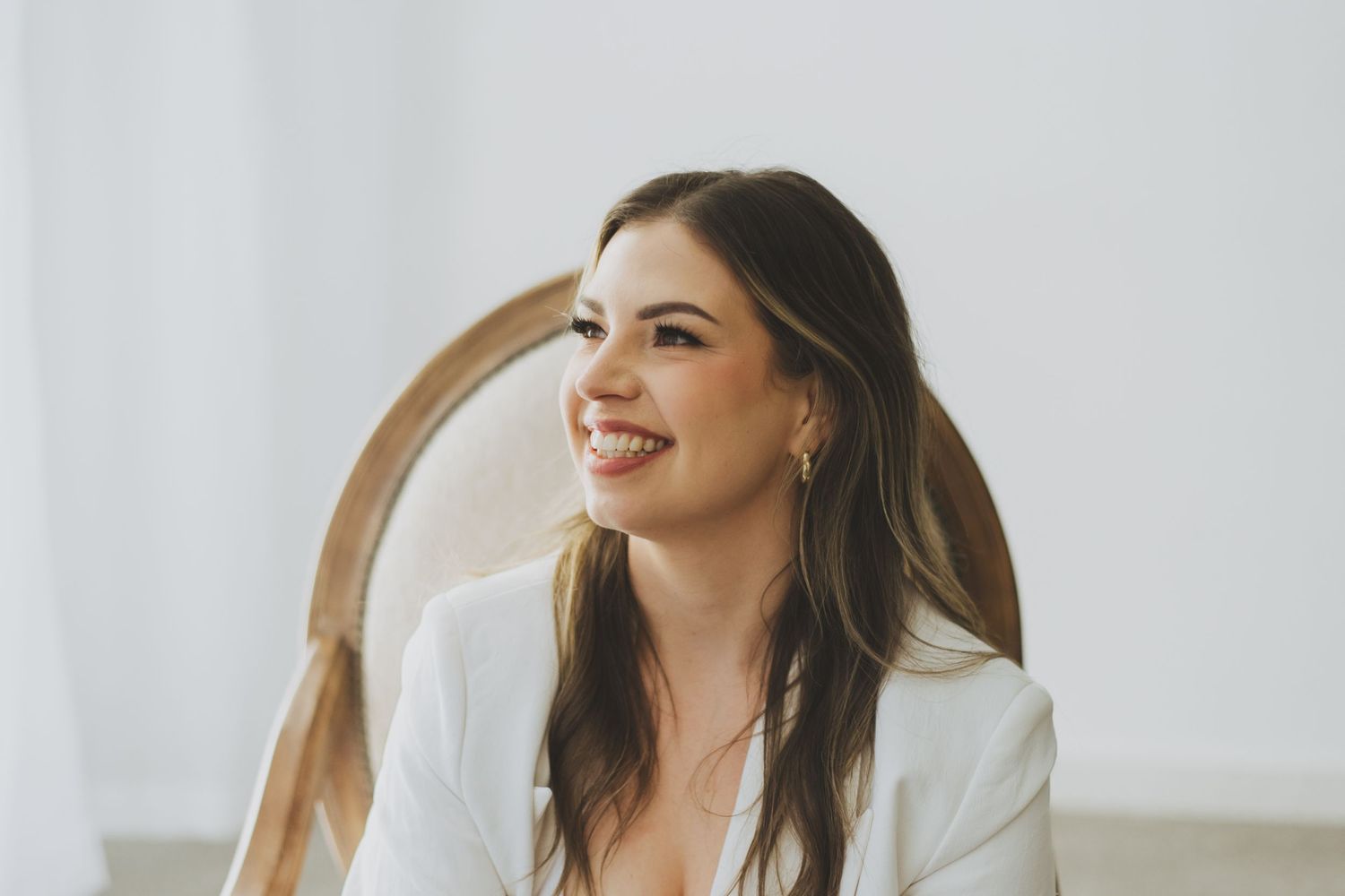 A woman in a white linen blazer smiles while sitting in a wooden vintage chair against a bright background.