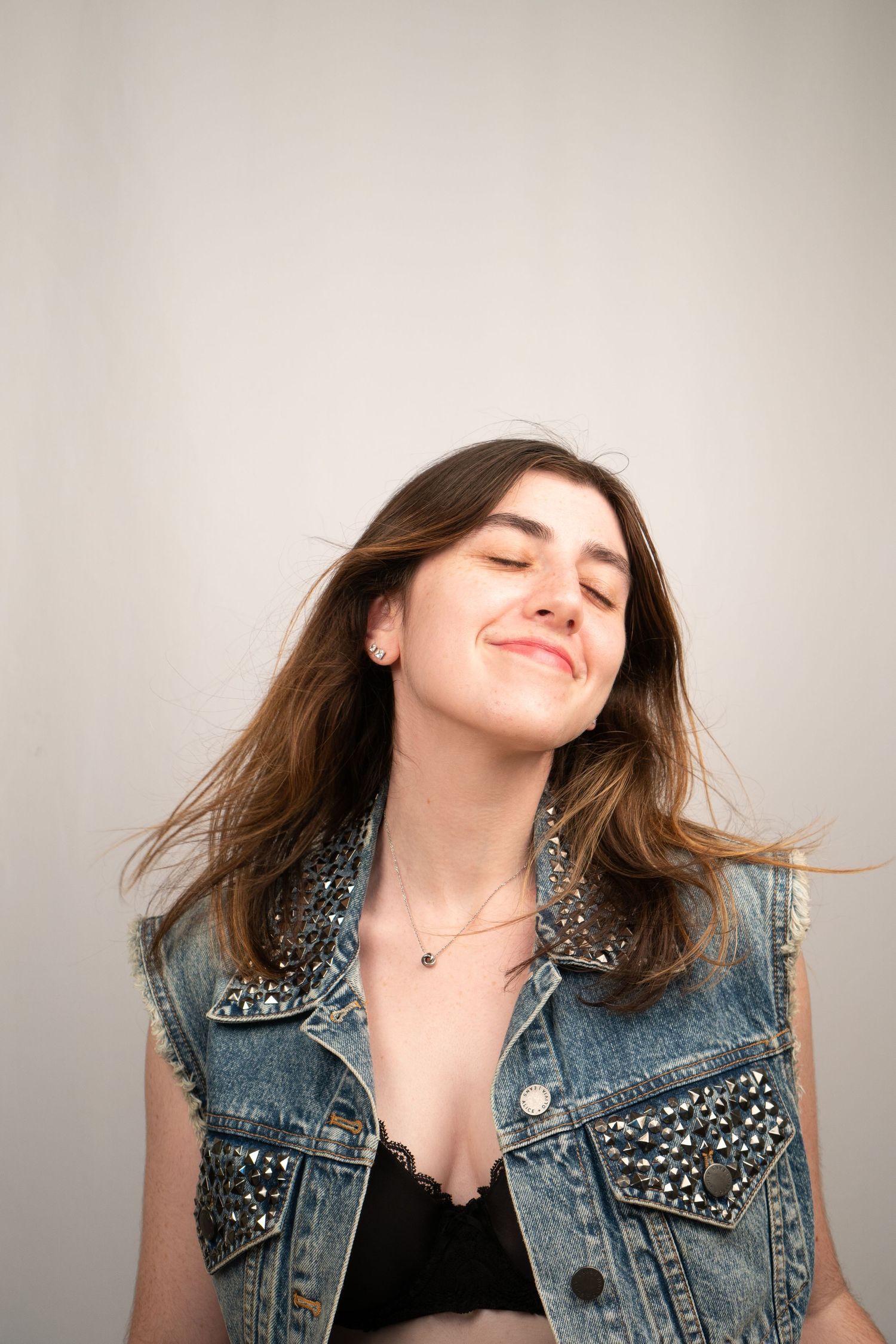 Woman with closed eyes wearing studded denim vest against light background.