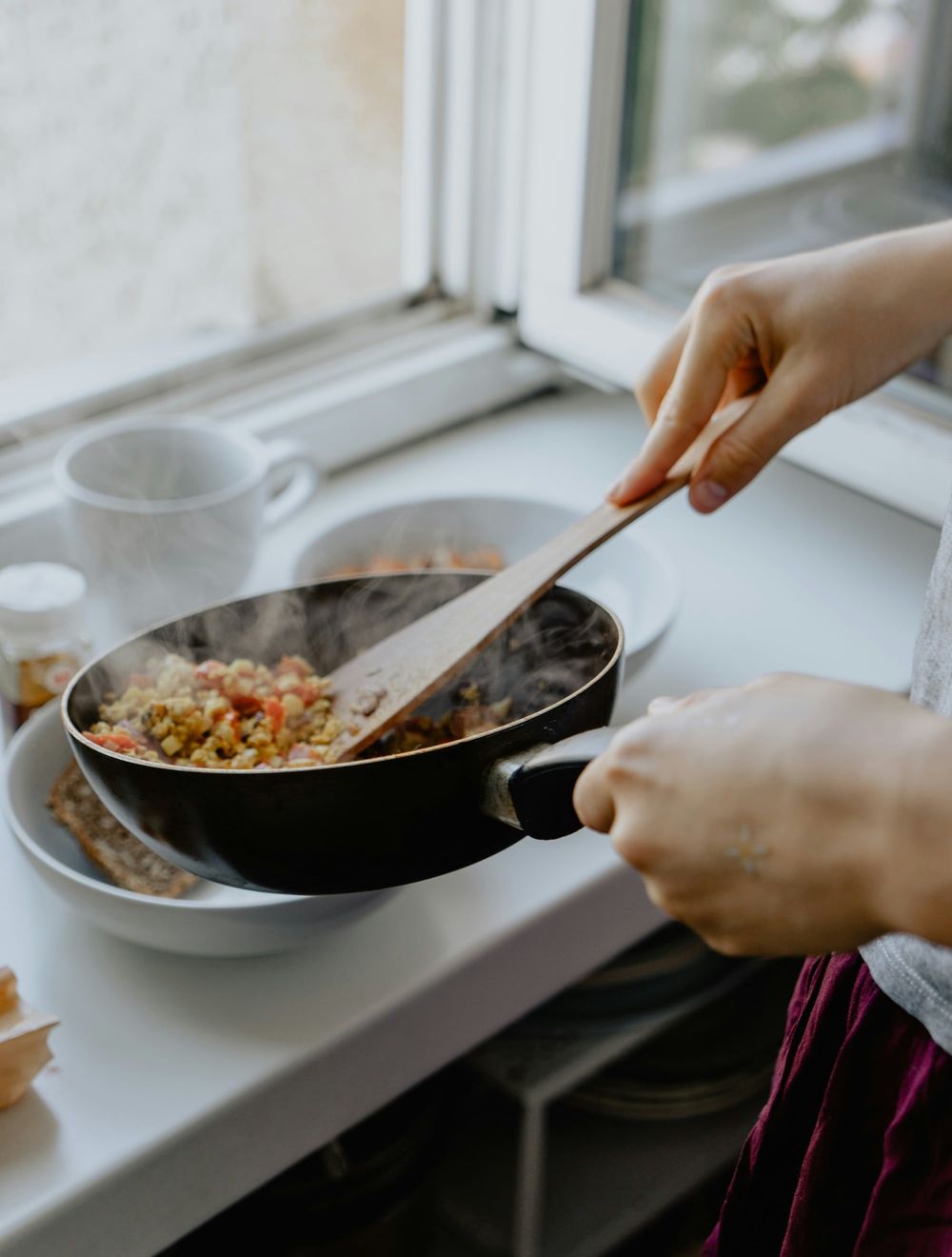 Person cooking and stirring food in a black pan by a window with natural lighting.