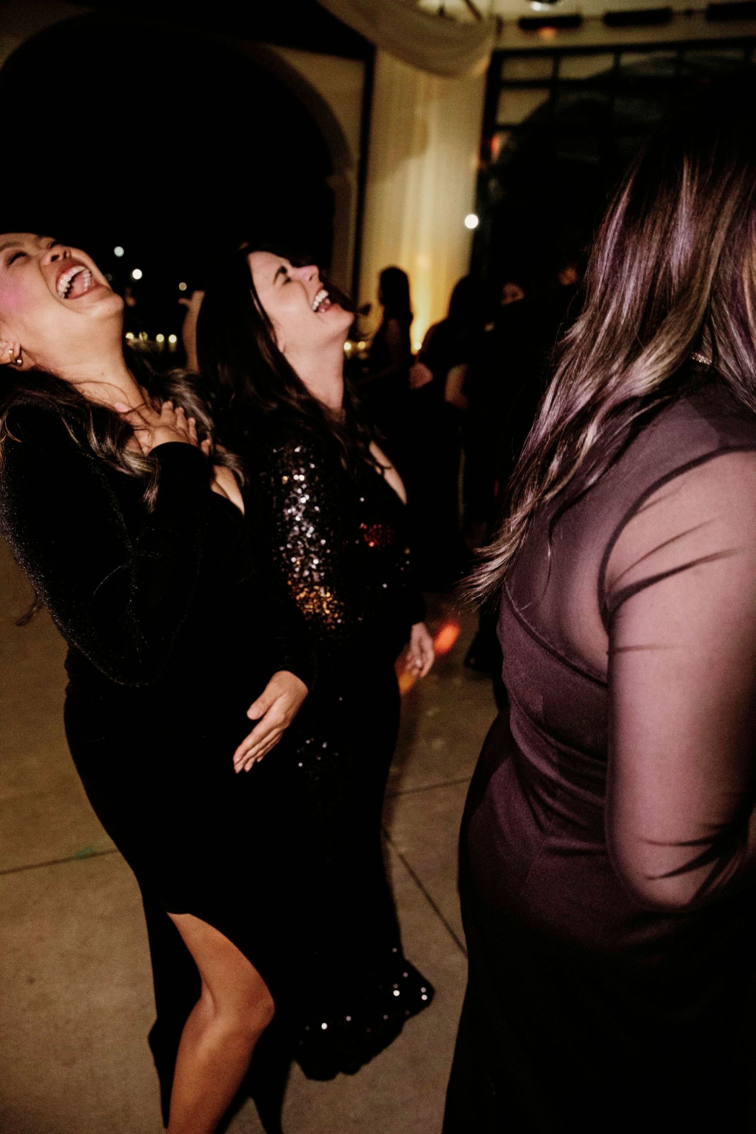 Party guests laugh and dance together at a celebration event wearing black and sequined formal dresses.