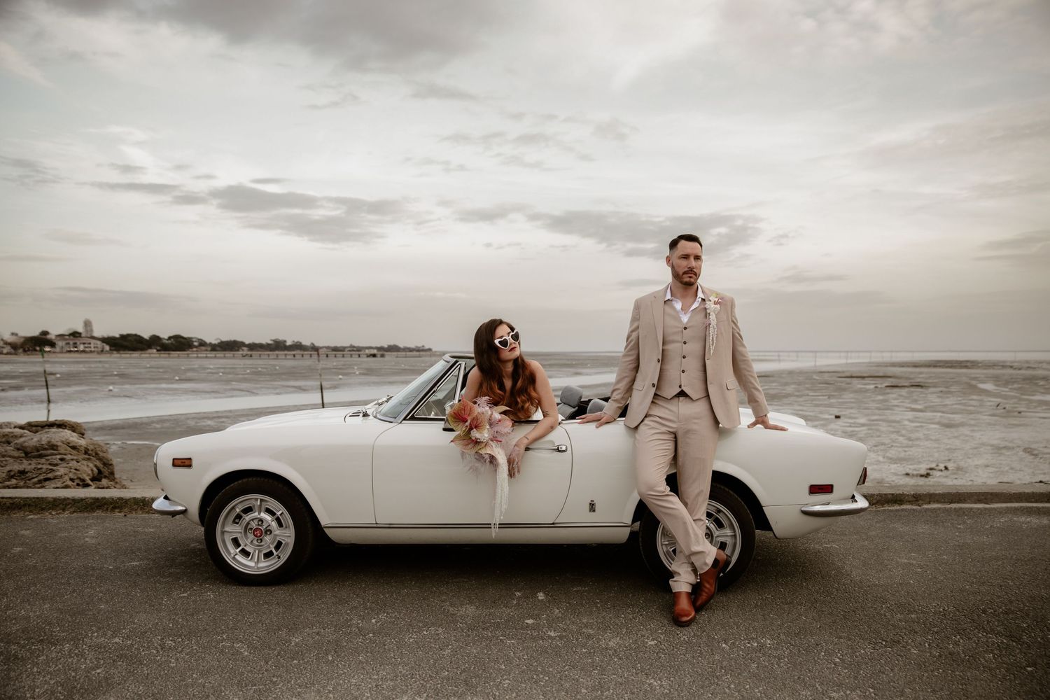 A classic white convertible car parked oceanside serves as a backdrop for stylish wedding photos.