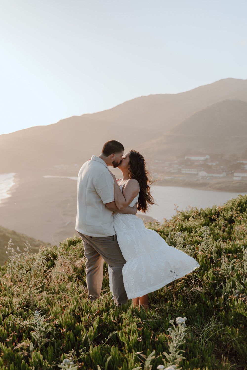 Couple embraces on hilltop during golden sunset with mountains and ocean in background.