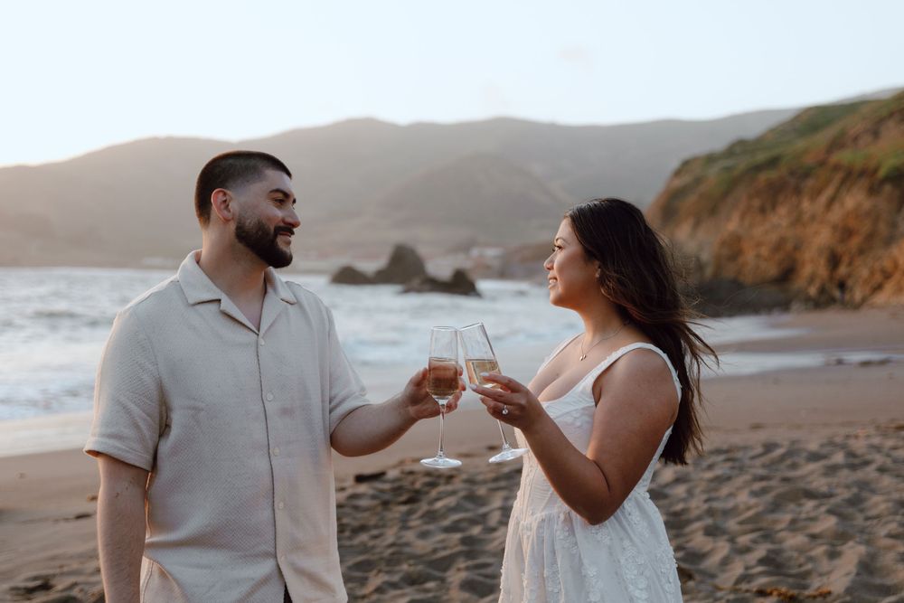A couple sharing a toast with wine glasses on a scenic beach at sunset with coastal cliffs in the background.