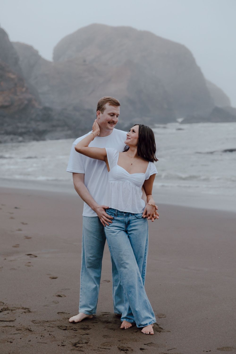 A couple in white tops and jeans share a romantic moment on a foggy beach with dramatic cliffs in the background.