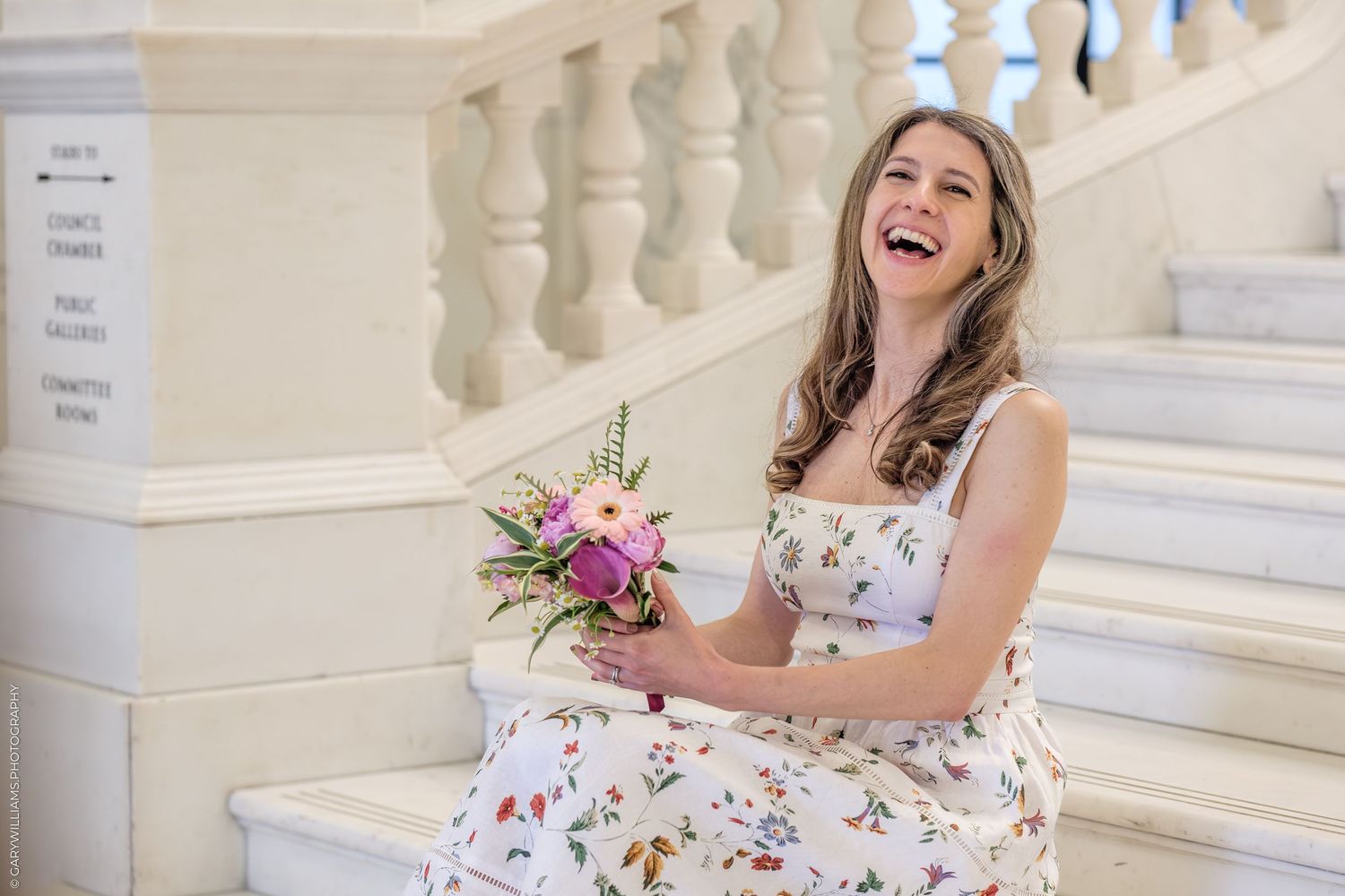 Someone in a floral dress laughs joyfully while sitting on elegant white stone steps at an outdoor venue.
