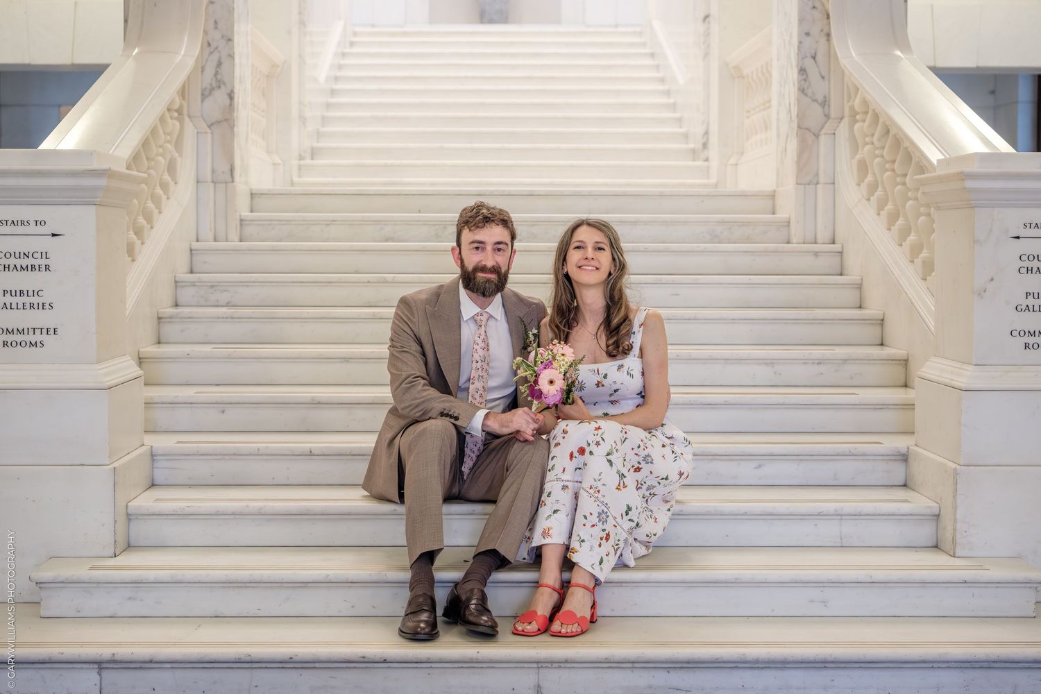 Couple sits together on elegant white marble staircase in coordinated formal attire for wedding photos or portraits.