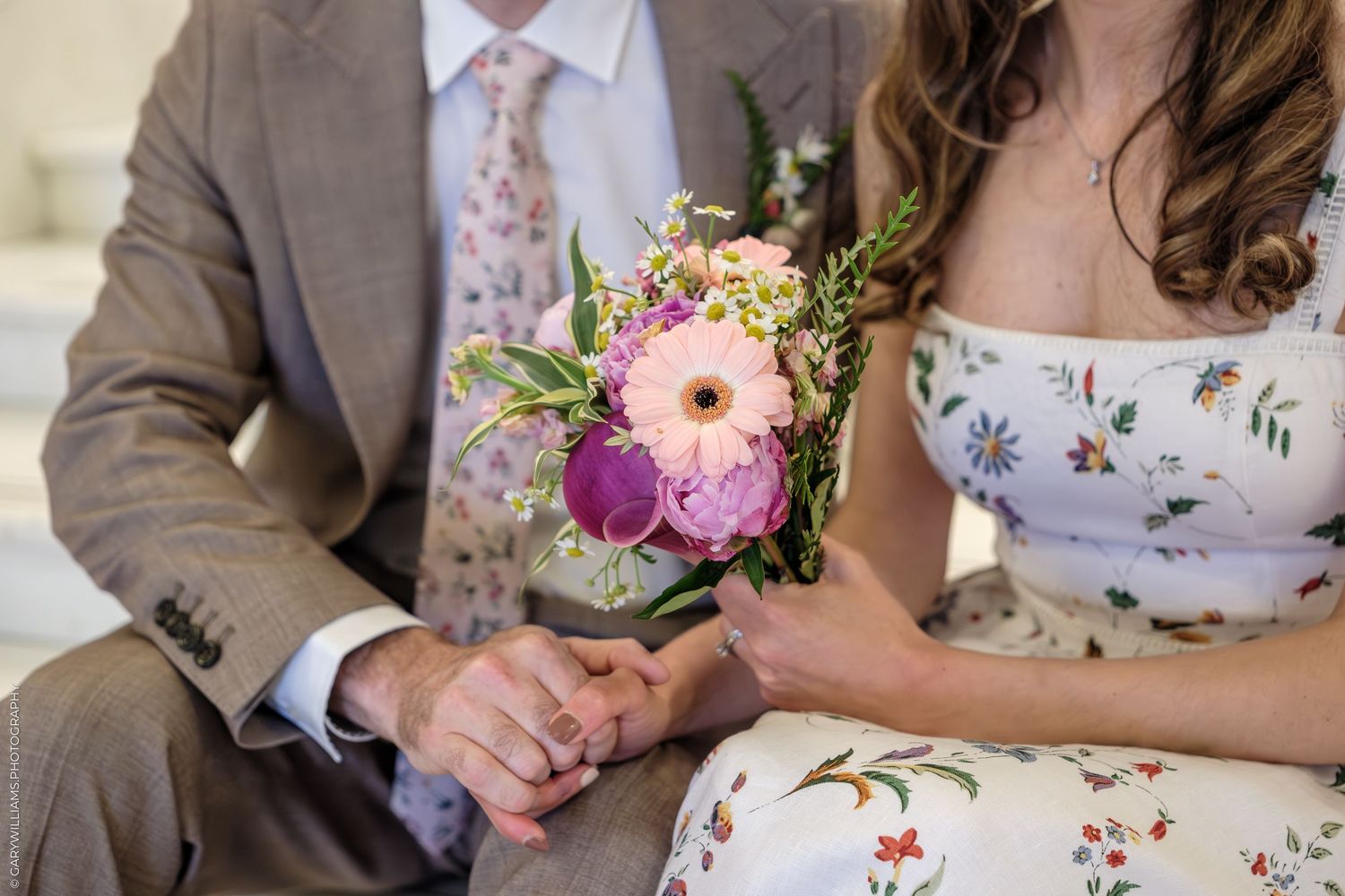 Close-up view of pink and purple wedding bouquet held against floral dress with tan suit visible in background.