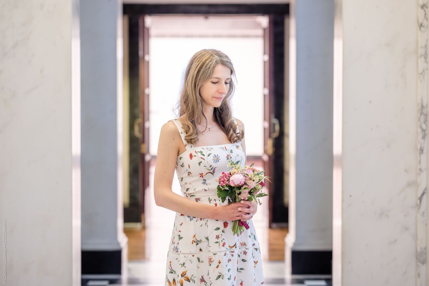 A bride in a floral print dress poses along a white column-lined walkway during a photoshoot.