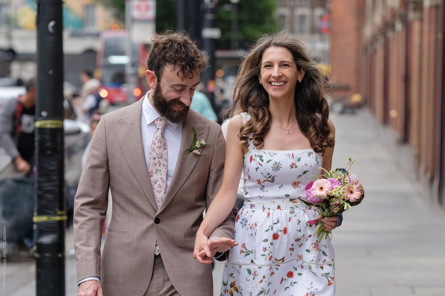 A couple walks down a city sidewalk, with the bride wearing a floral dress and holding a bouquet while walking with her partner.