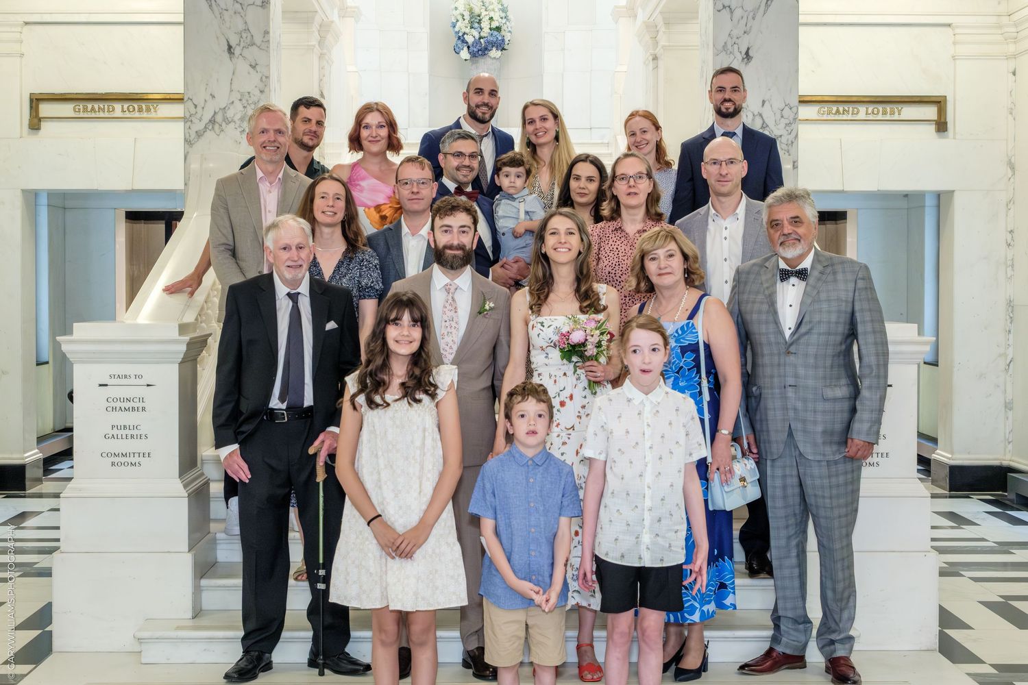 A formal family group stands gathered together on white marble steps inside an elegant building interior.