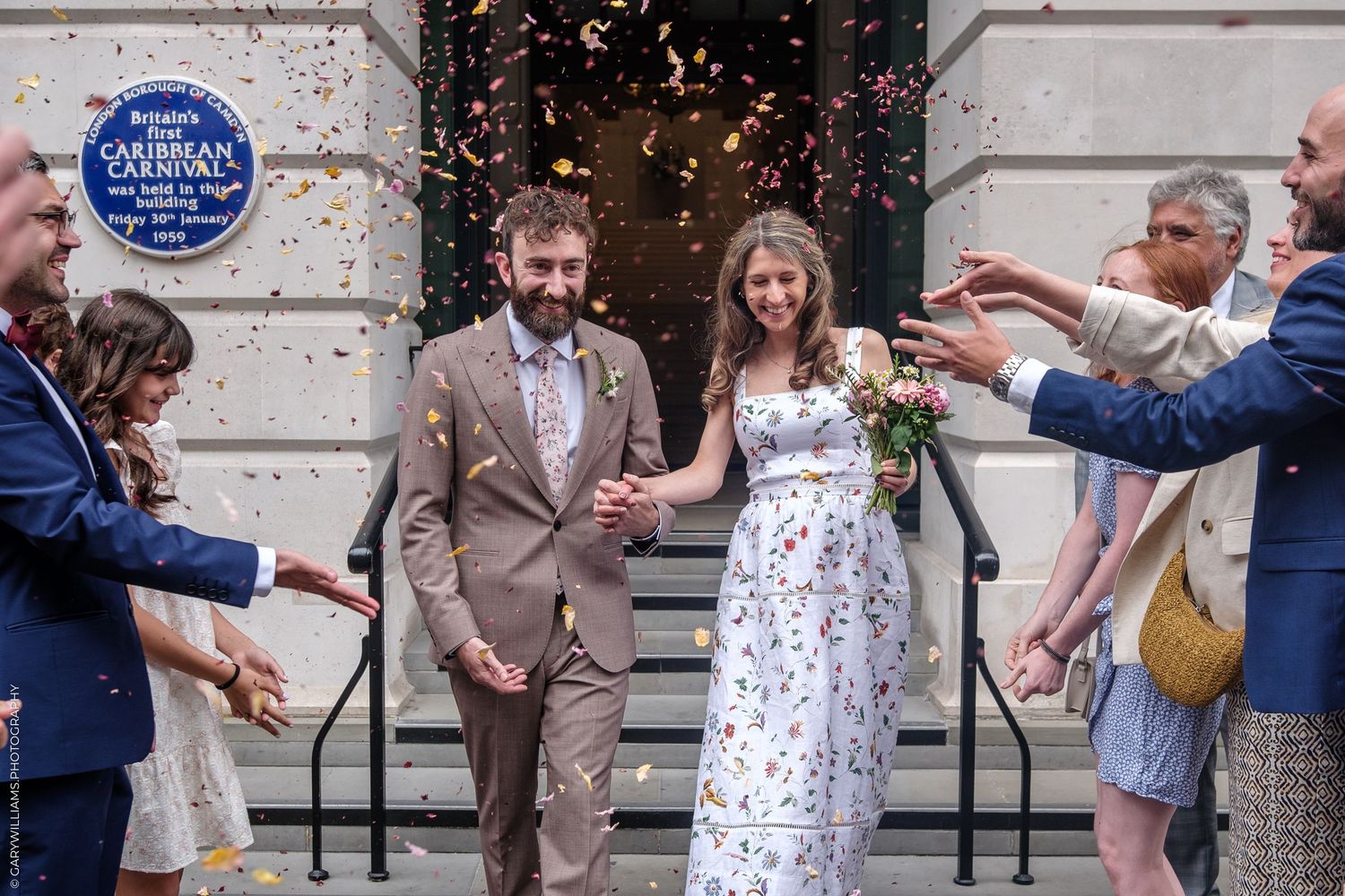 Newlyweds exit down steps while guests joyfully throw confetti during celebration outside building with blue plaque.
