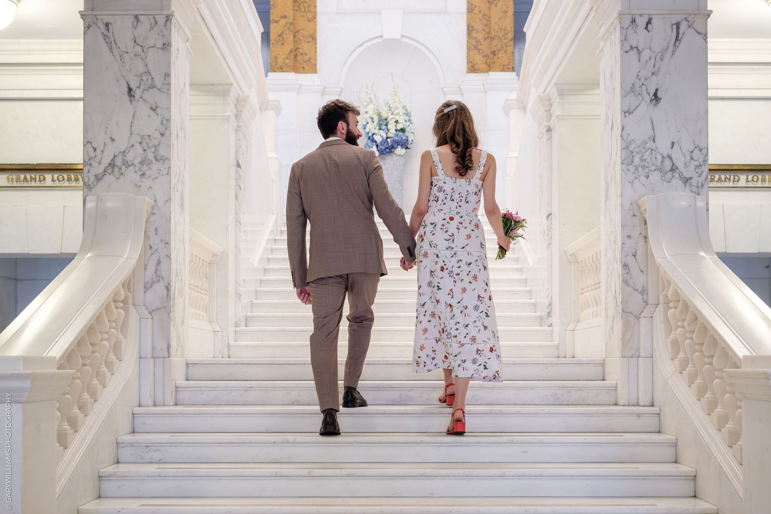 A couple in formal attire walks up a white marble staircase holding hands in a bright elegant interior setting.