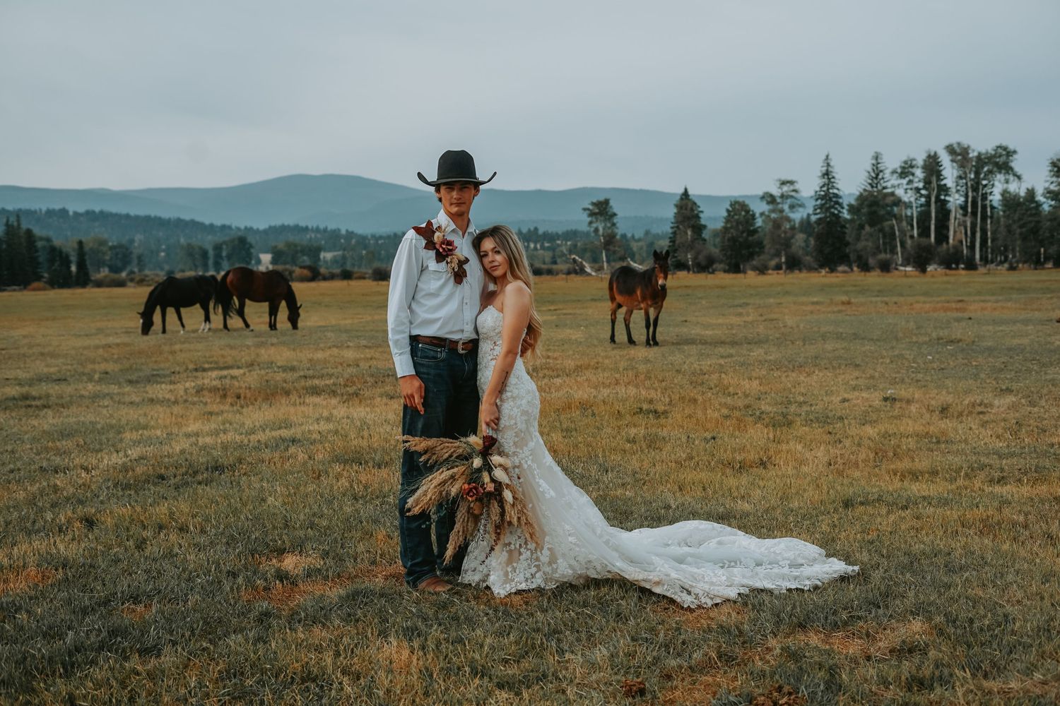 A couple poses for their wedding photo with horses grazing in the distance.