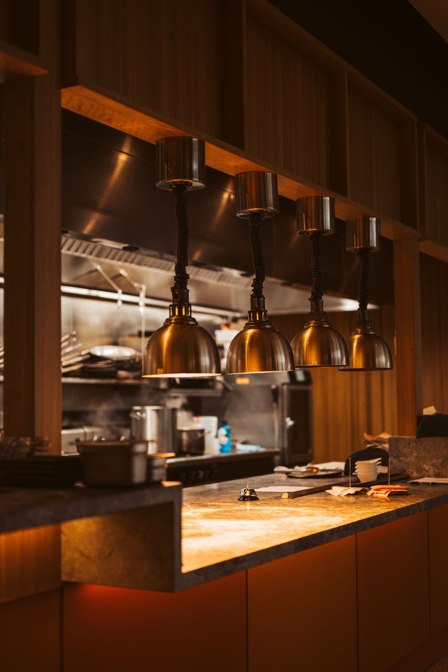 Modern restaurant kitchen pass-through with warm copper pendant lights illuminating a wooden counter and open kitchen in the background
