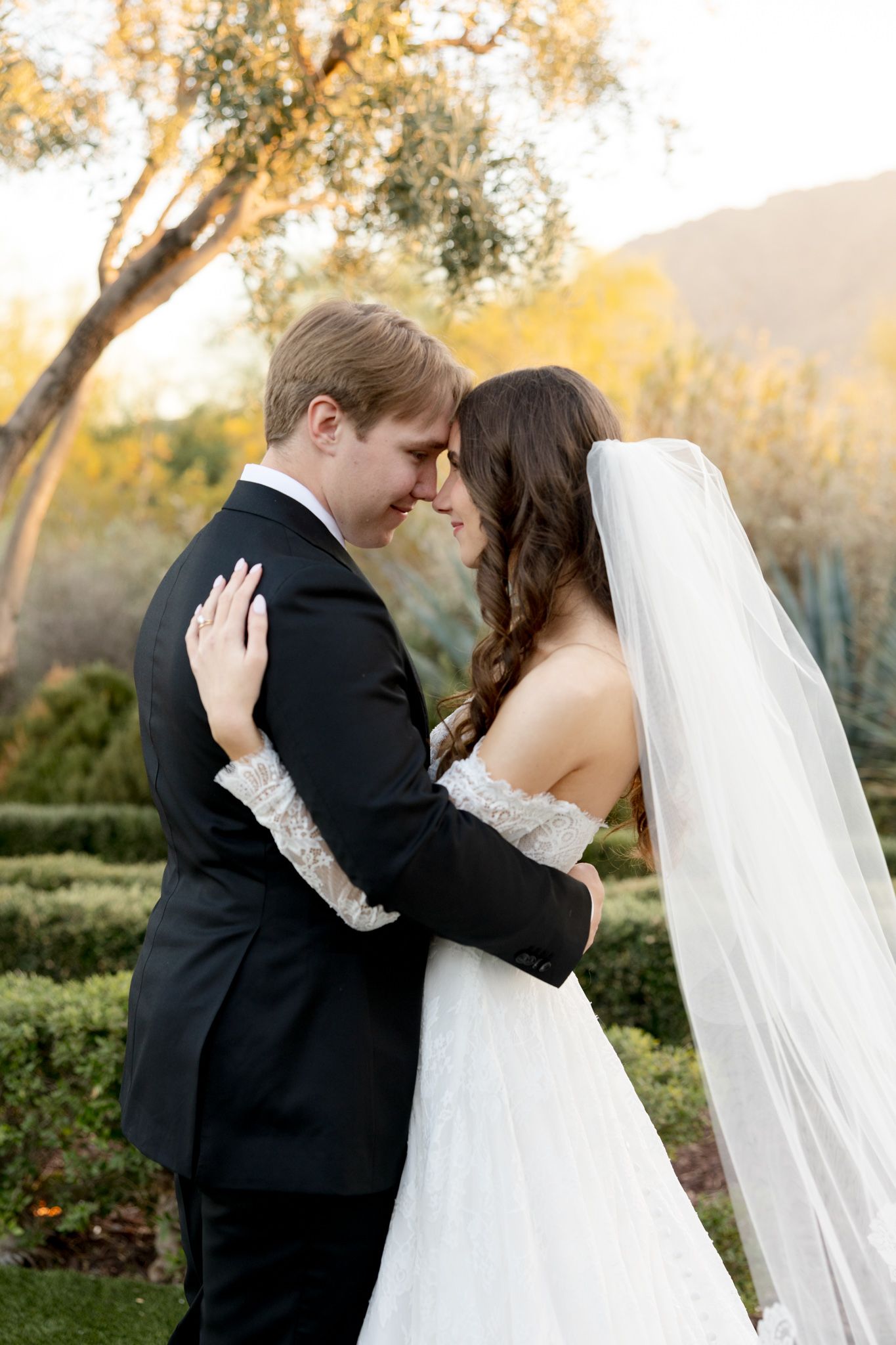 Romantic wedding couple embraces outdoors with flowing white veil and autumn foliage in background during golden hour.
