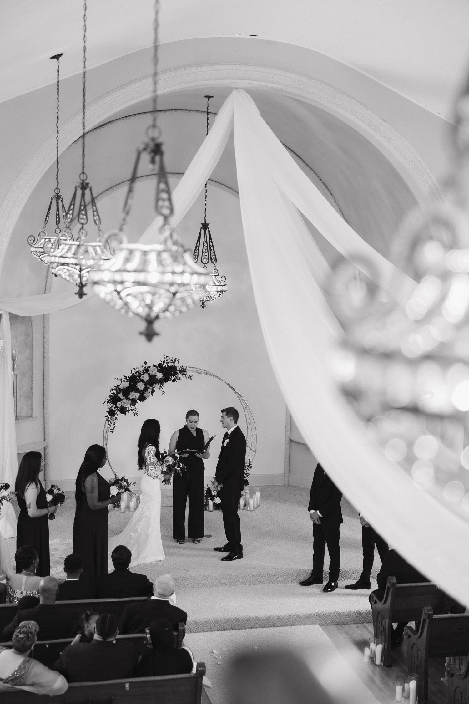 A black and white wedding ceremony takes place under an elegant arched chapel interior with ornate chandeliers.