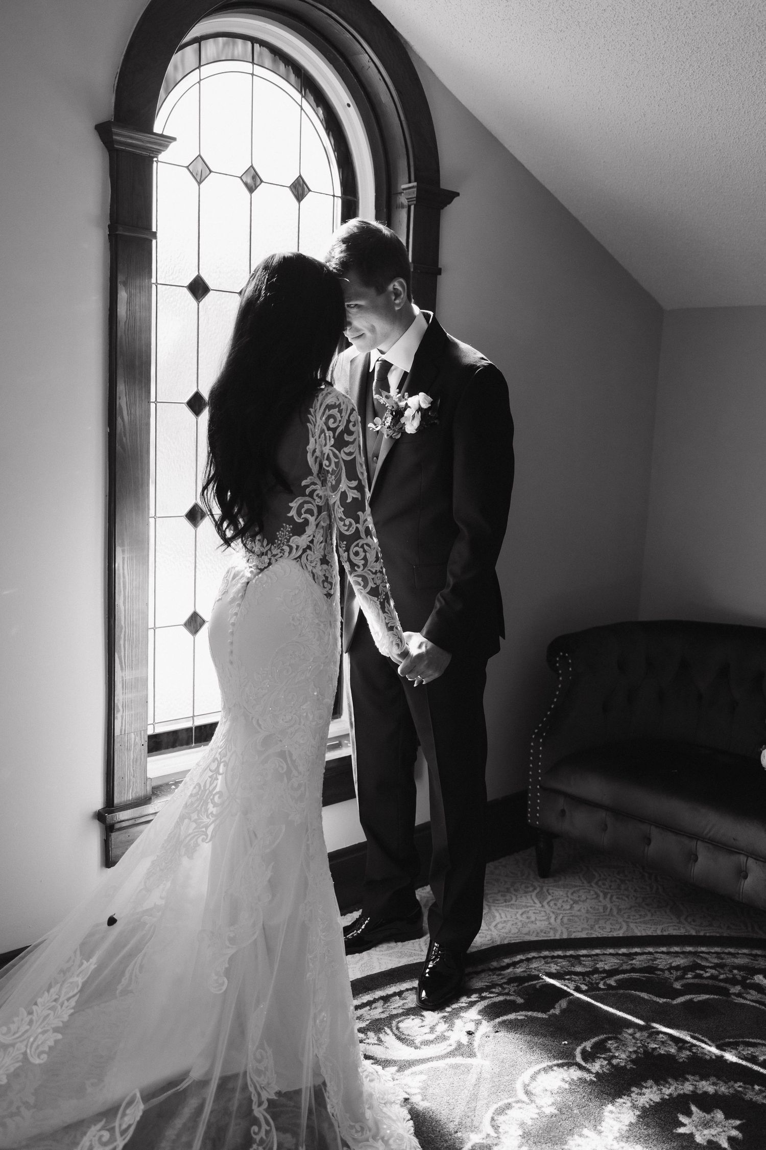 A black and white portrait of newlyweds sharing an intimate moment by an ornate arched window.