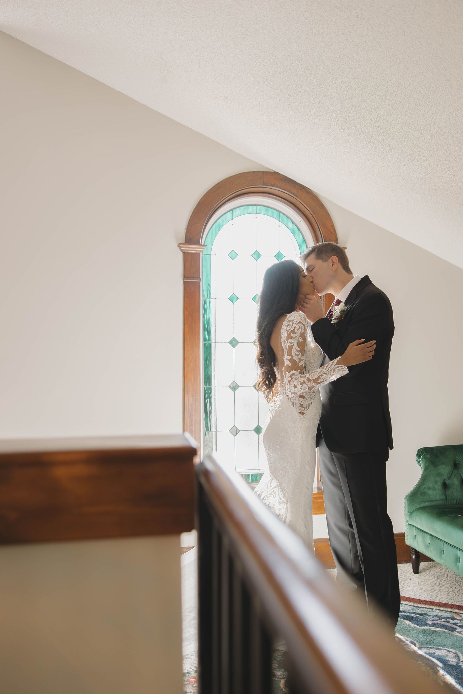 A romantic wedding photo of a couple embracing by an arched stained glass window in an elegant interior.