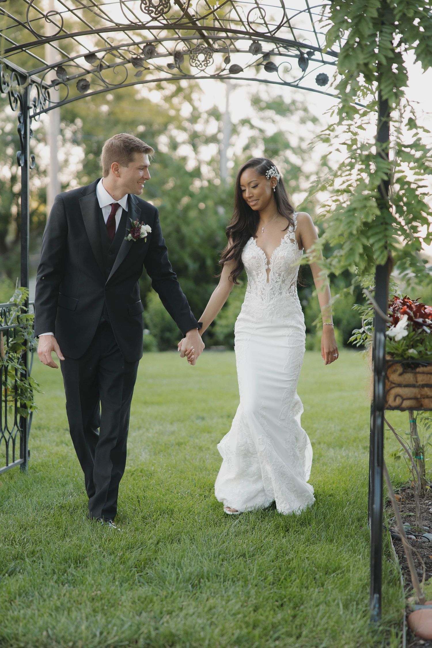 A couple walks under a garden arbor surrounded by lush greenery during a wedding celebration.
