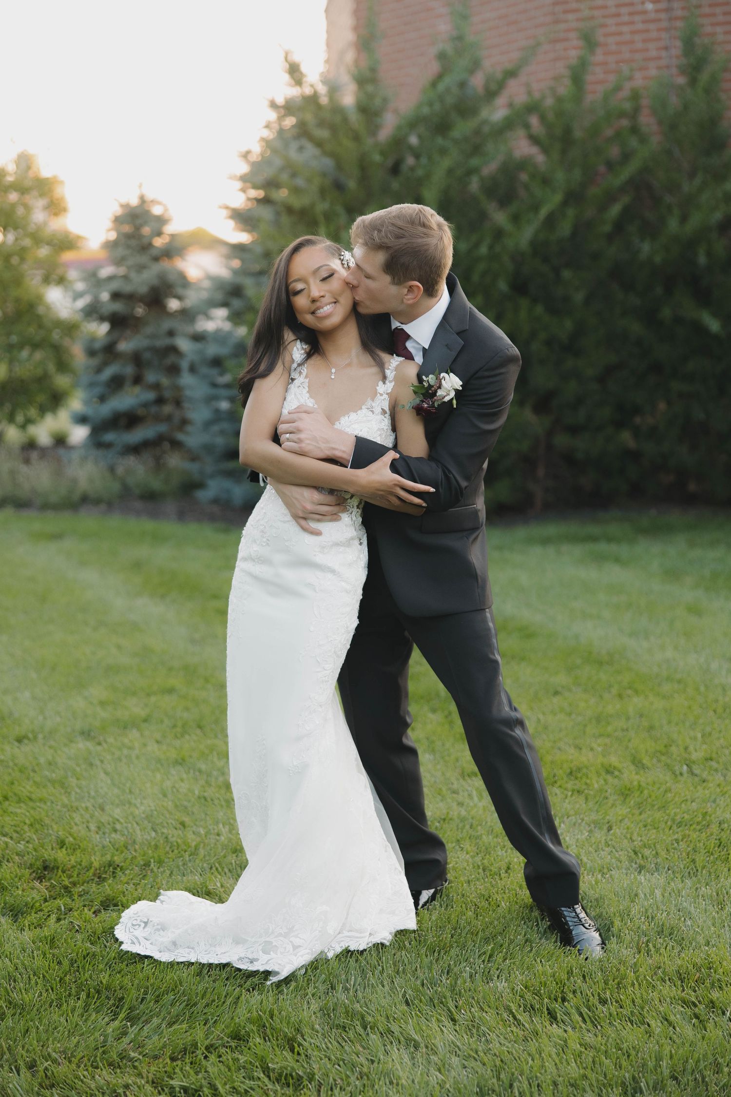 Wedding couple dances playfully on a green lawn with brick building in the background.