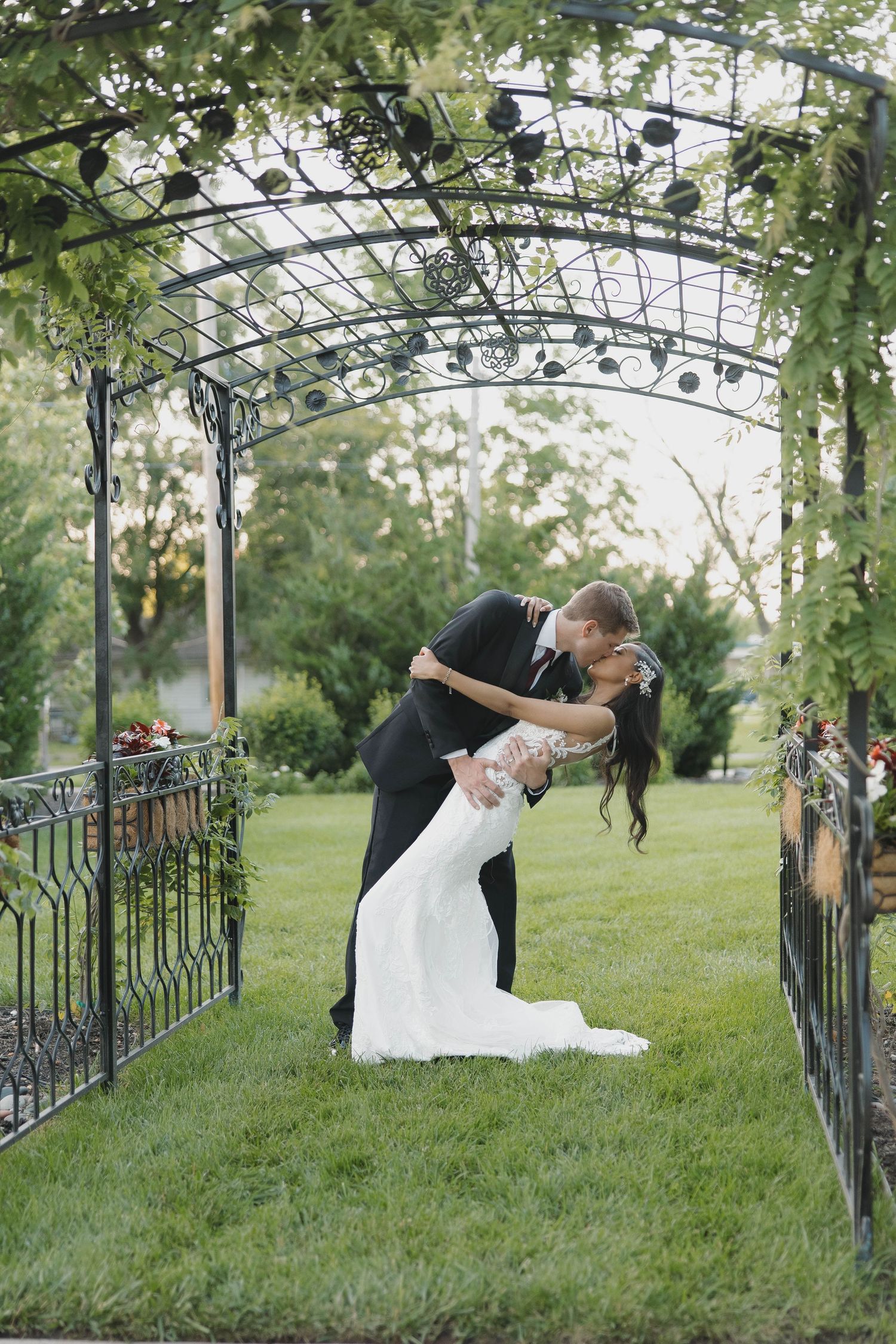 A romantic moment under a vine-covered garden archway as a couple embraces in formal wedding attire on a green lawn.