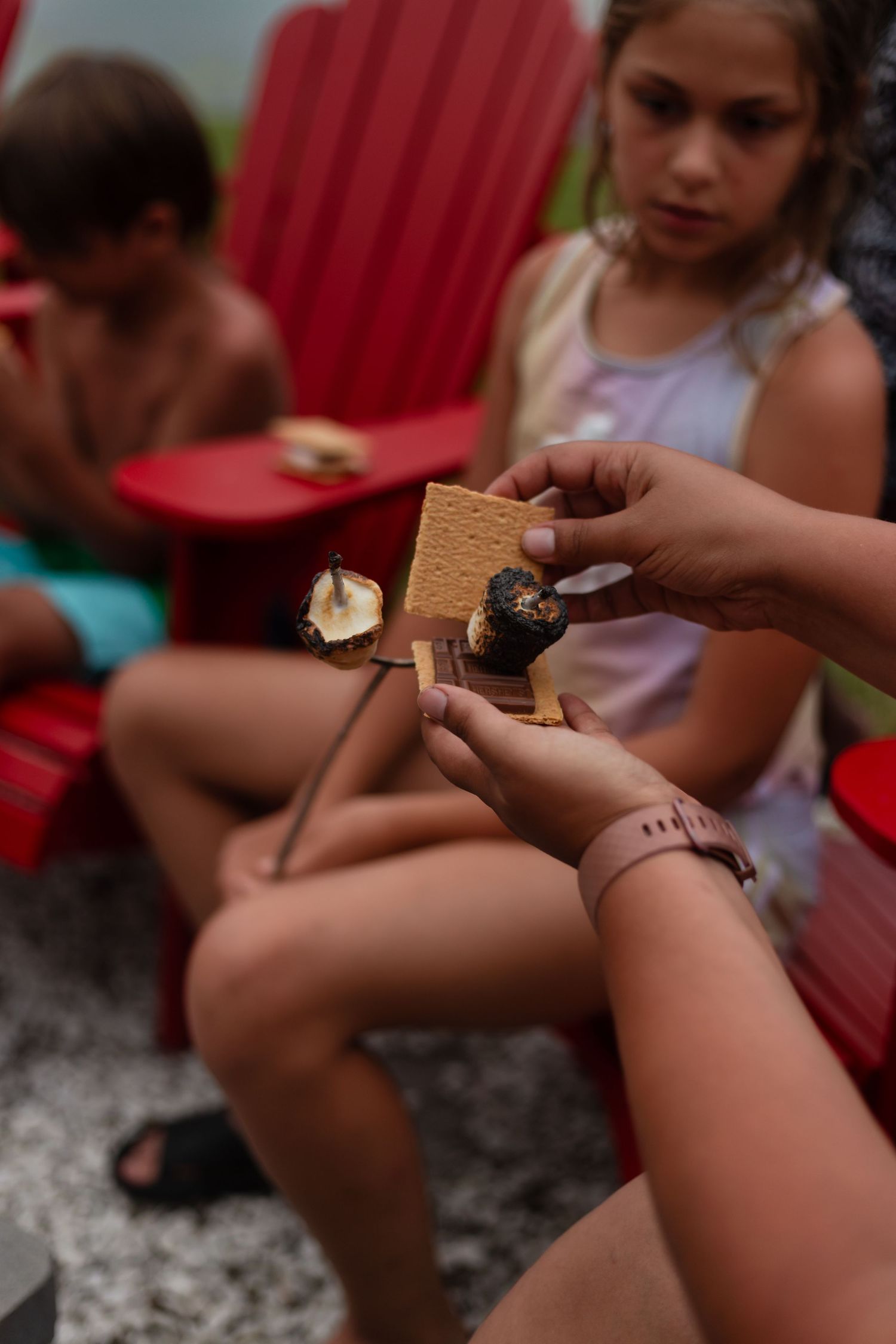 Several hands are assembling classic s'mores treats outdoors with graham crackers, marshmallows and chocolate bars.