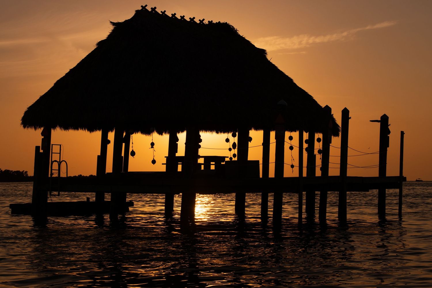 Silhouettes of seaside structures stand against a warm orange sunset sky reflecting on calm ocean waters.