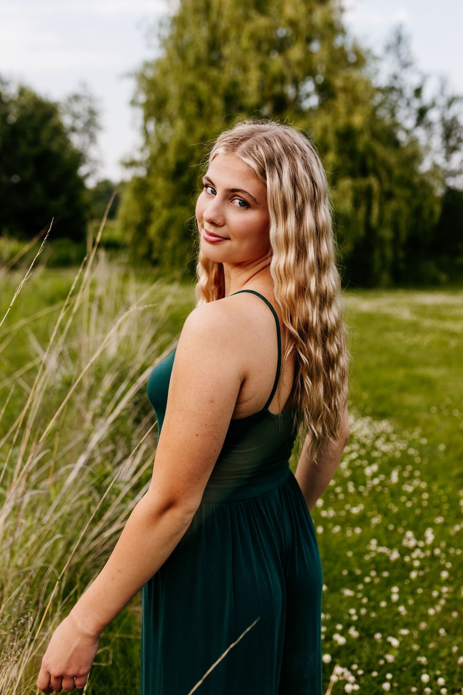 A person in an elegant dark green dress poses in a summer meadow with tall grass and wildflowers.