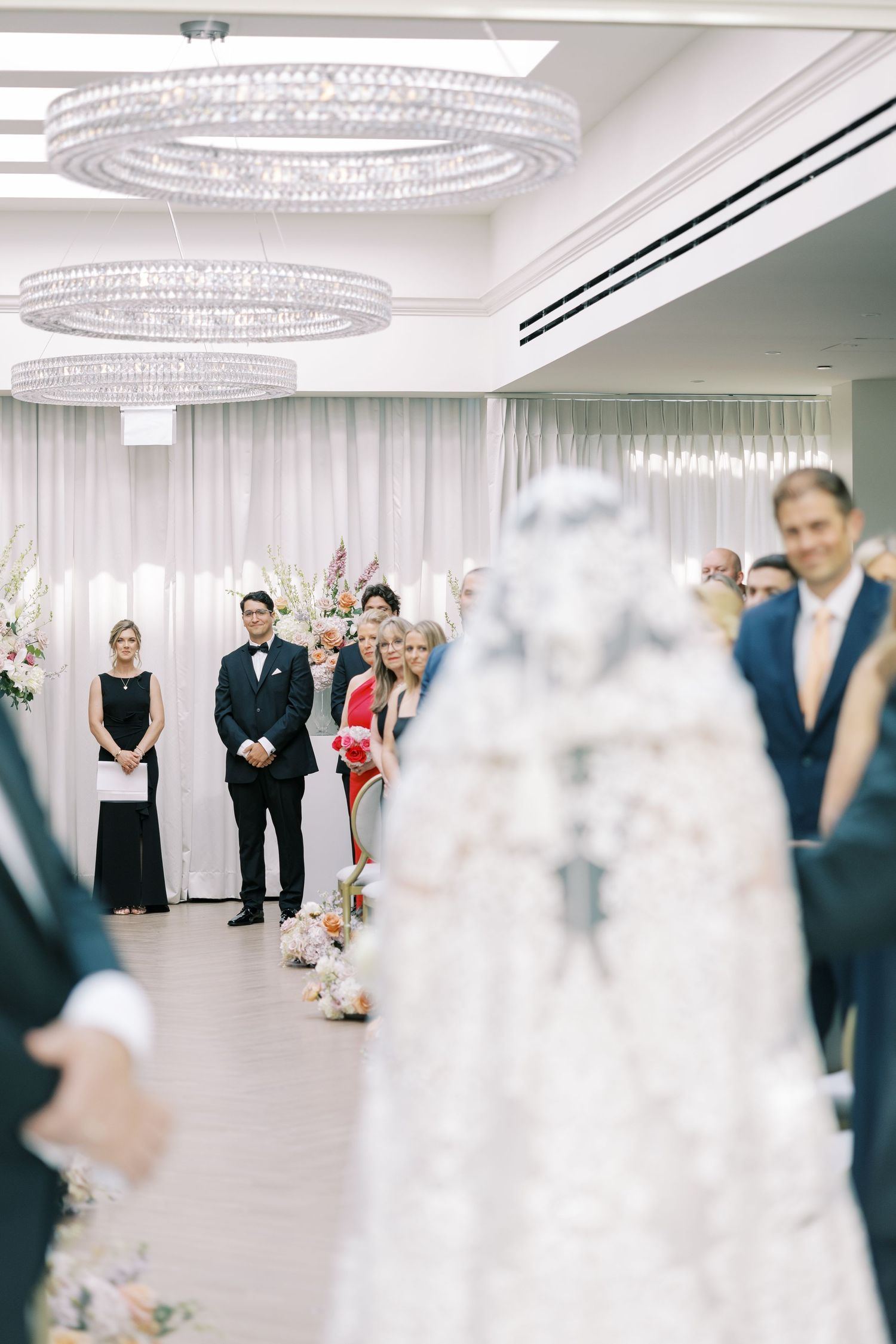 Wedding ceremony in a modern venue with crystal chandeliers and white sheer curtains as guests look on during the service.