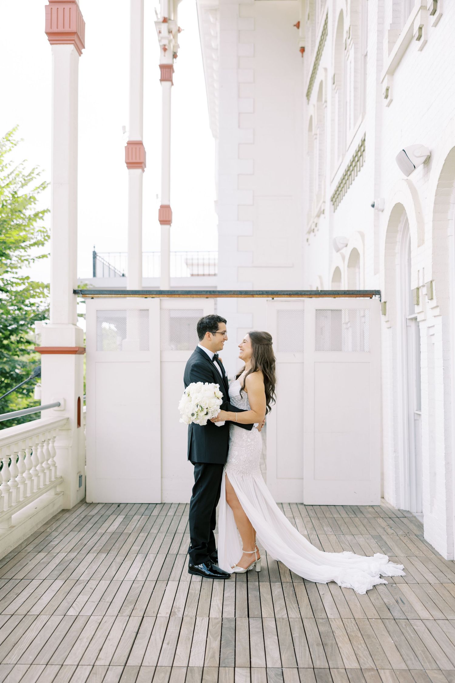 Elegant wedding couple embracing on a white columned balcony with wooden deck flooring and white floral bouquet.