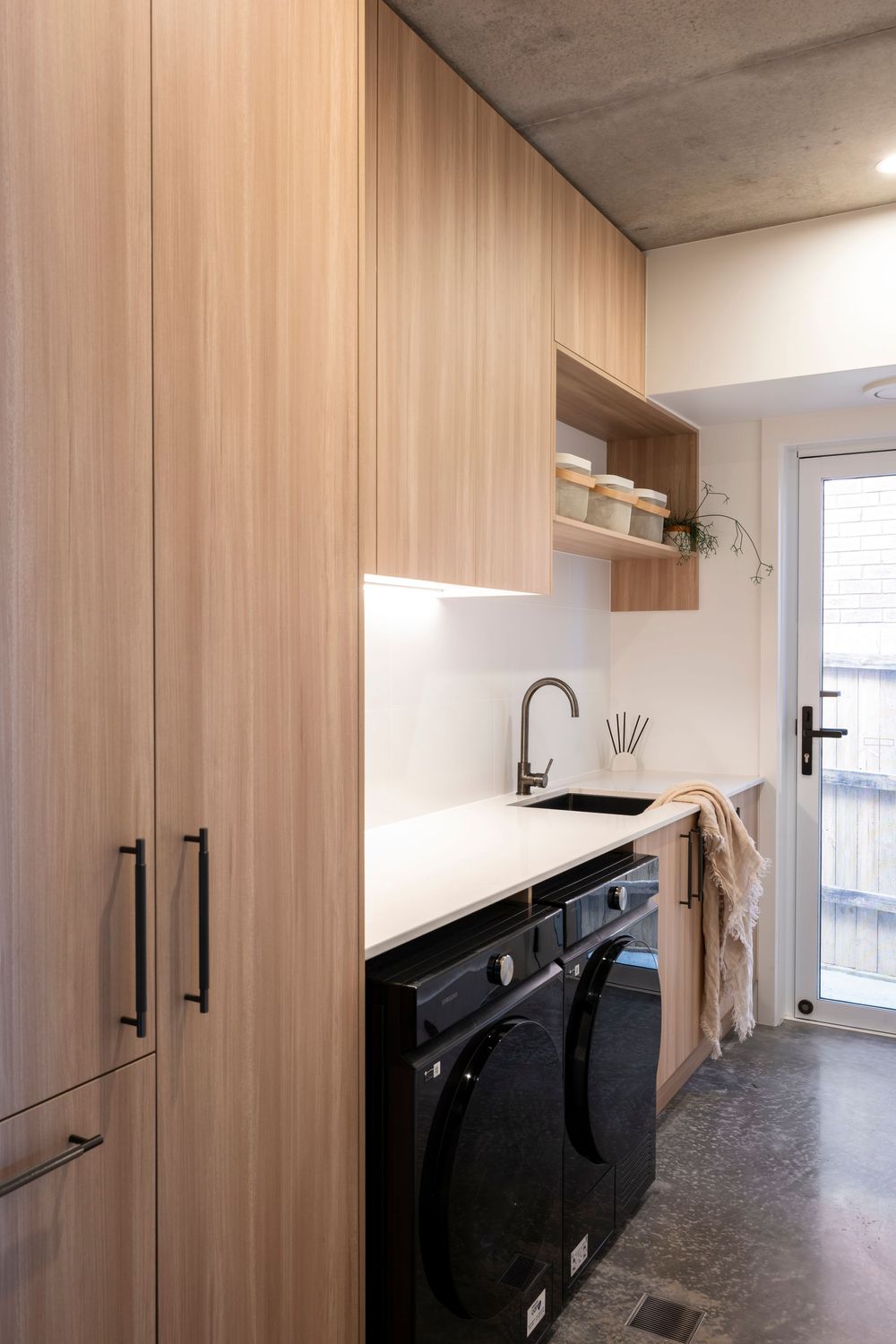 Modern laundry room with light wood cabinetry, black appliances, white countertops and matte black hardware details.