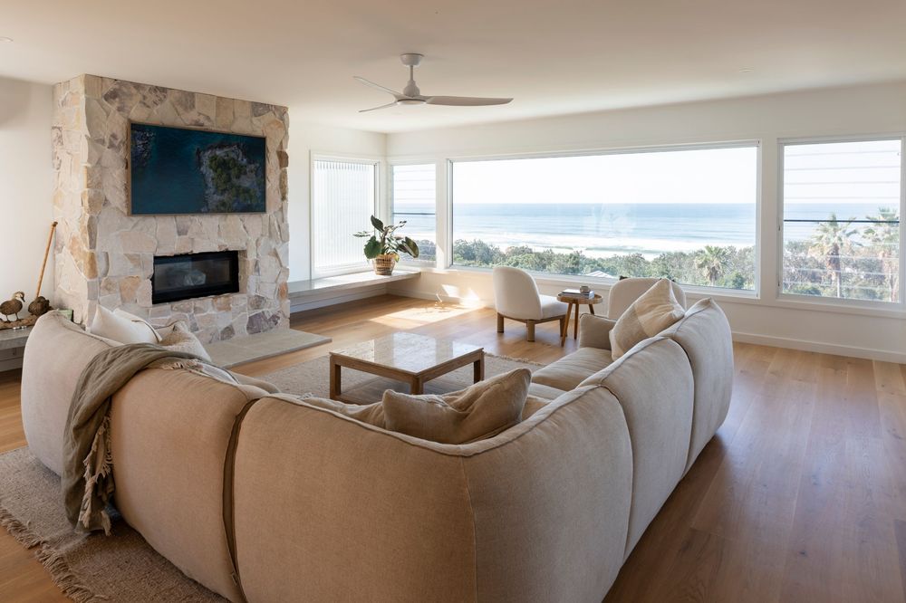 Modern coastal living room with beige sectional sofa, stone fireplace and panoramic ocean views through large windows.