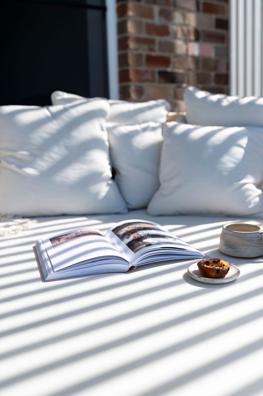 Sunlight casting dramatic venetian blind shadows across a white couch with pillows and an open book with coffee cup.