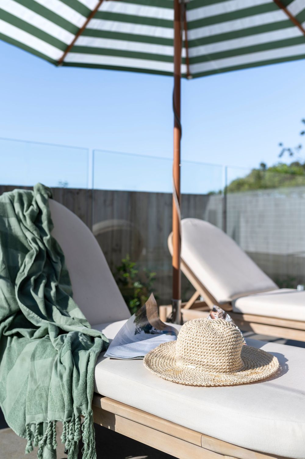 A serene patio setting with a striped umbrella, white lounge cushions, green throw, and straw hat under bright sunlight.