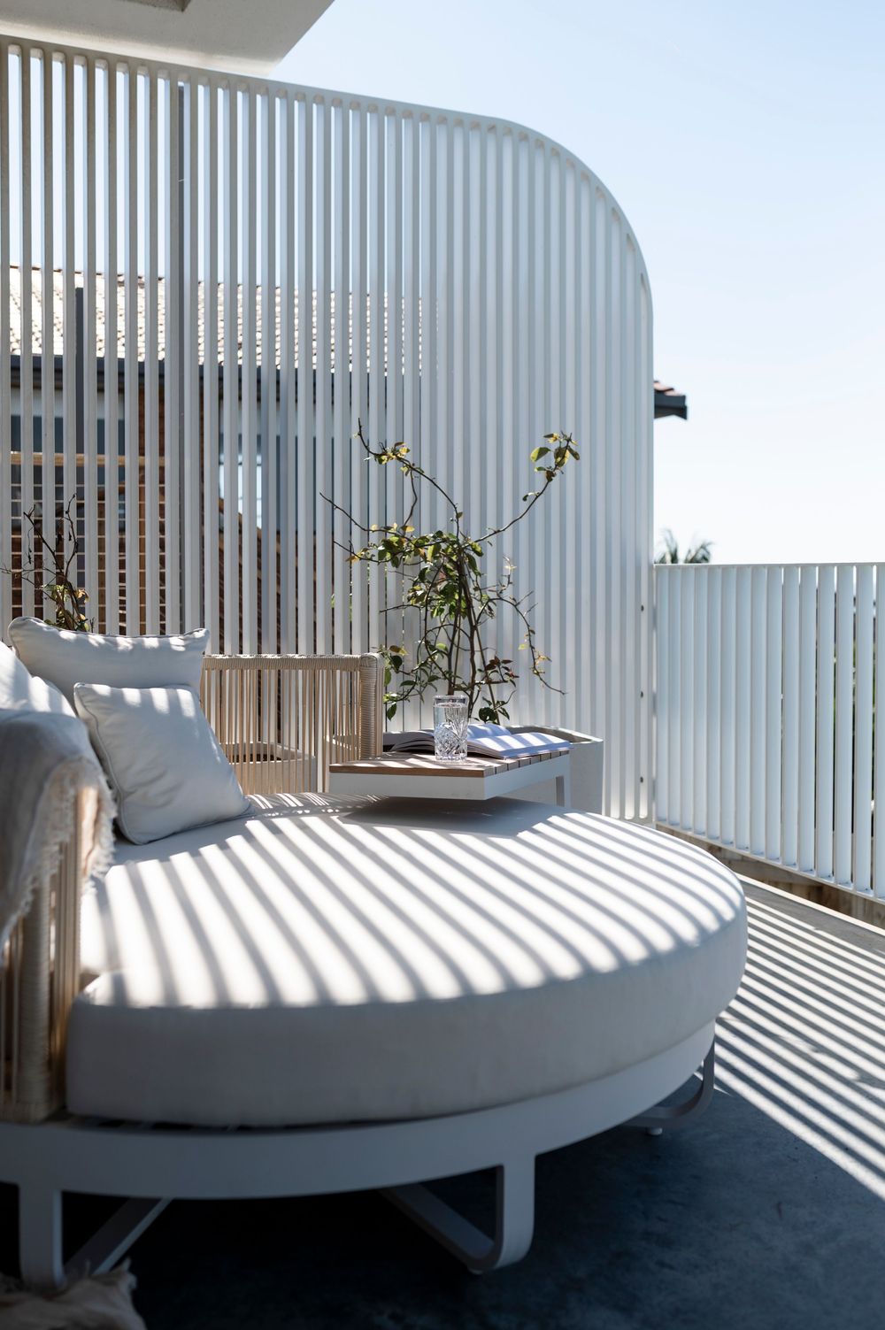 Curved white daybed with pillows on modern balcony featuring vertical slat privacy screens casting dramatic shadows.