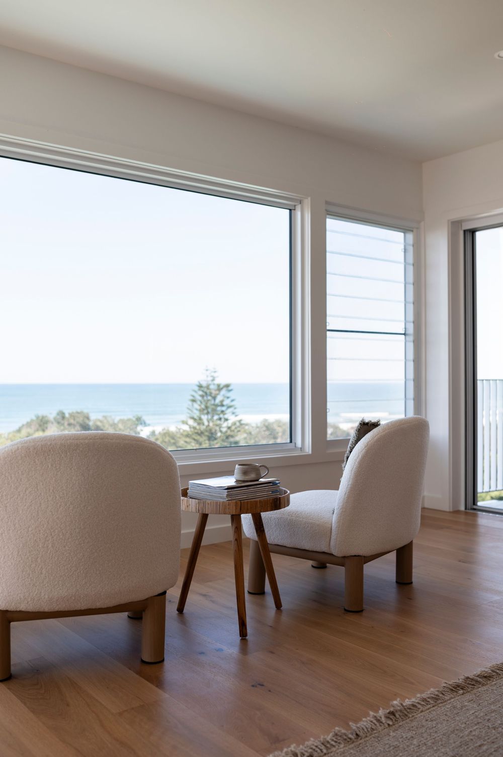 Modern coastal living room features two white boucle chairs and wooden side table with ocean view through large windows.