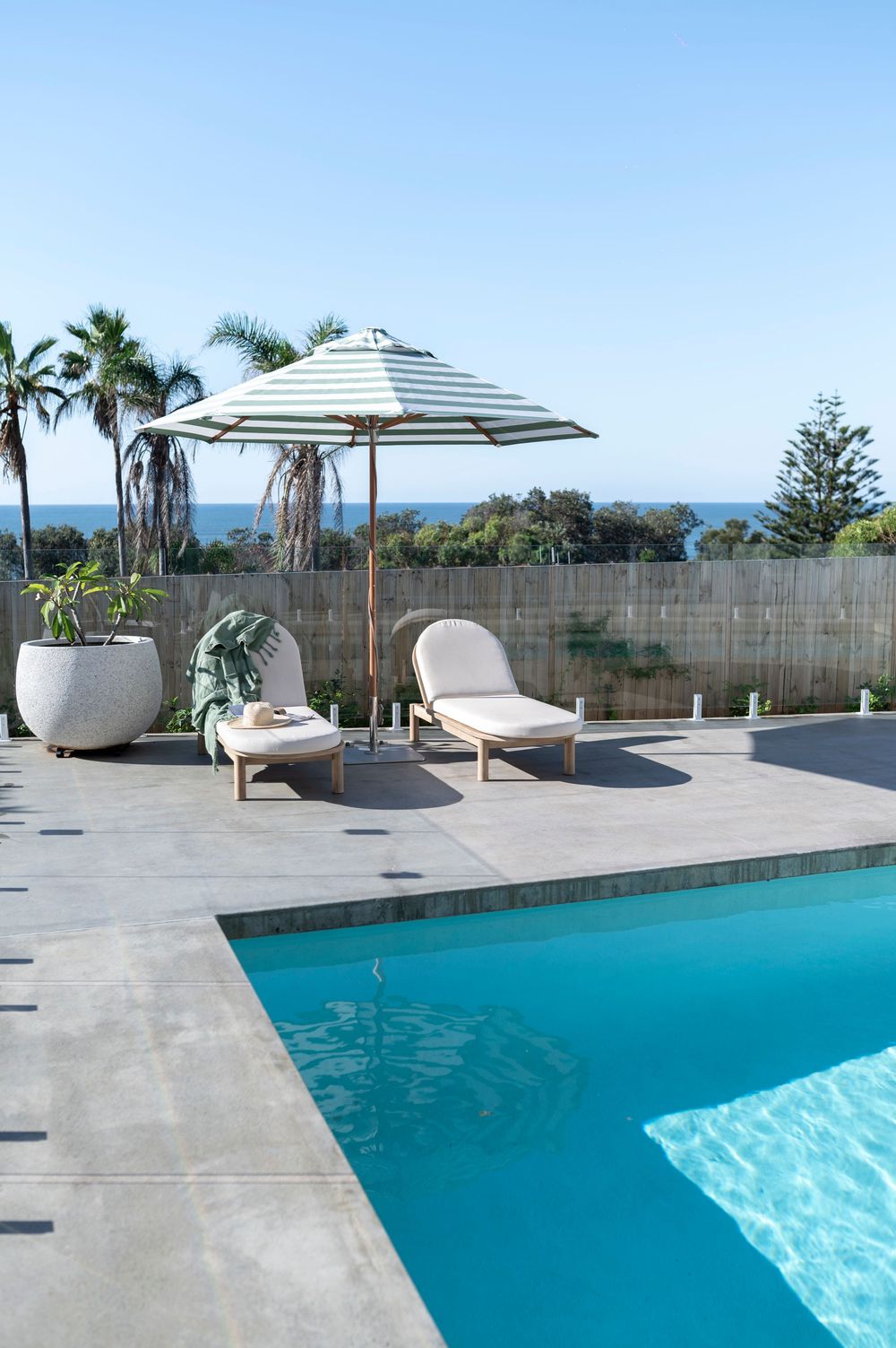 Modern poolside lounge area with striped umbrella and chaise loungers overlooking palm trees and ocean view.