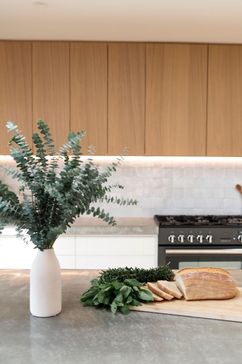 Modern kitchen counter with eucalyptus in white vase, fresh bread and herbs on cutting board beside black stove.