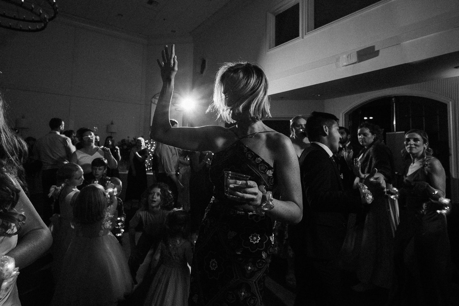 A crowd dances at a wedding reception while someone holds up their arm against the dark atmospheric lighting.