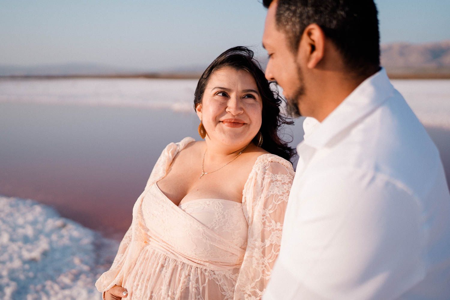 A couple embraces on salt flats at sunset while wearing white and pink romantic outfits.