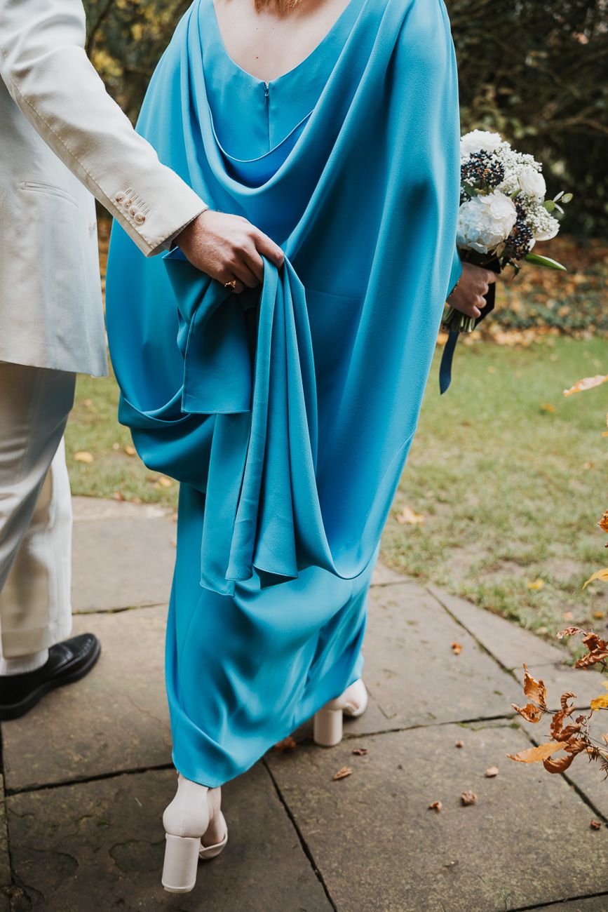 Close up of turquoise flowing dress and white shoes at an outdoor wedding with bouquet and suit visible.